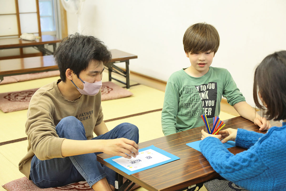 集会所の和室に並べられた低い長机に向かうボランティアの大学生とノルウェーから来日して一年のお子さん。