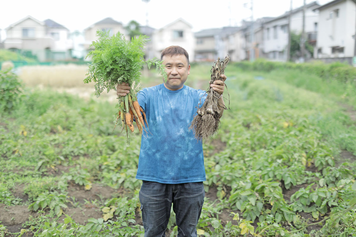 畑で、収穫した人参とにんにくをそれぞれの手に持って見せている男性。藍染めのようなTシャツを着ている。