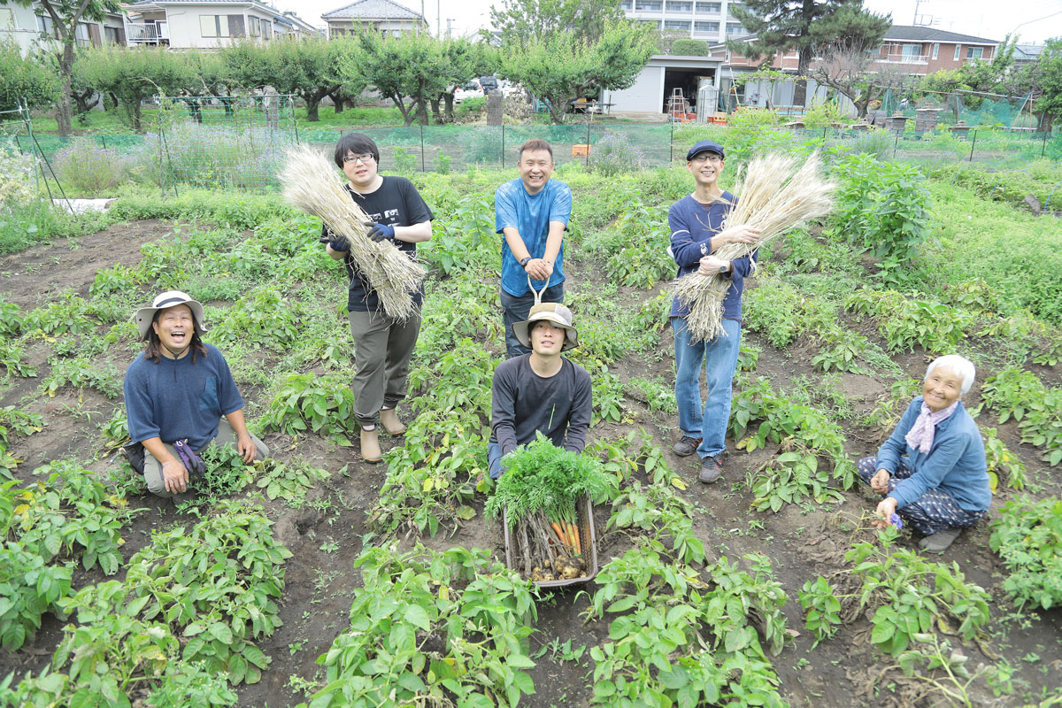 みんなで畑の畝の合間に並んで集合写真。収穫した麦や人参などの野菜を手にしている。