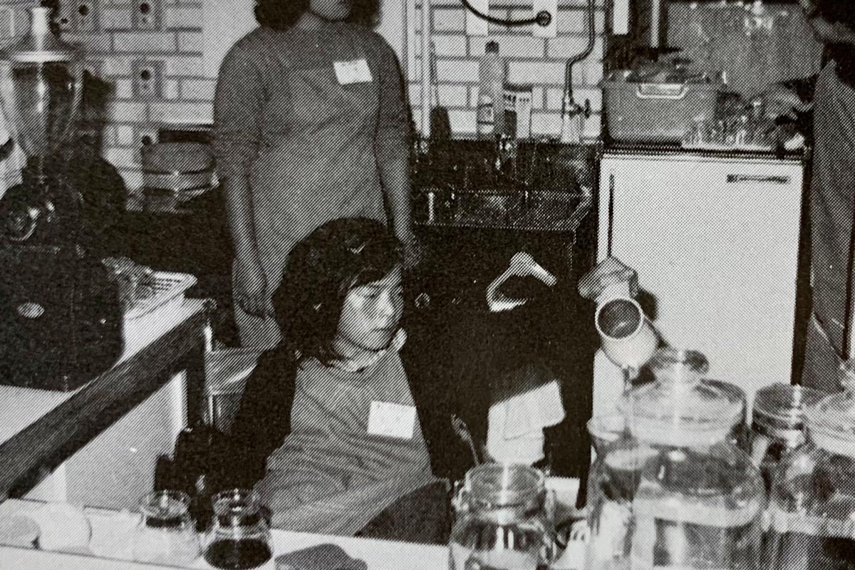 カフェの厨房で車いすに座った若い女性が、ほうろうの注ぎ口が細いポットで、ドリッパーにお湯を注いでいる。