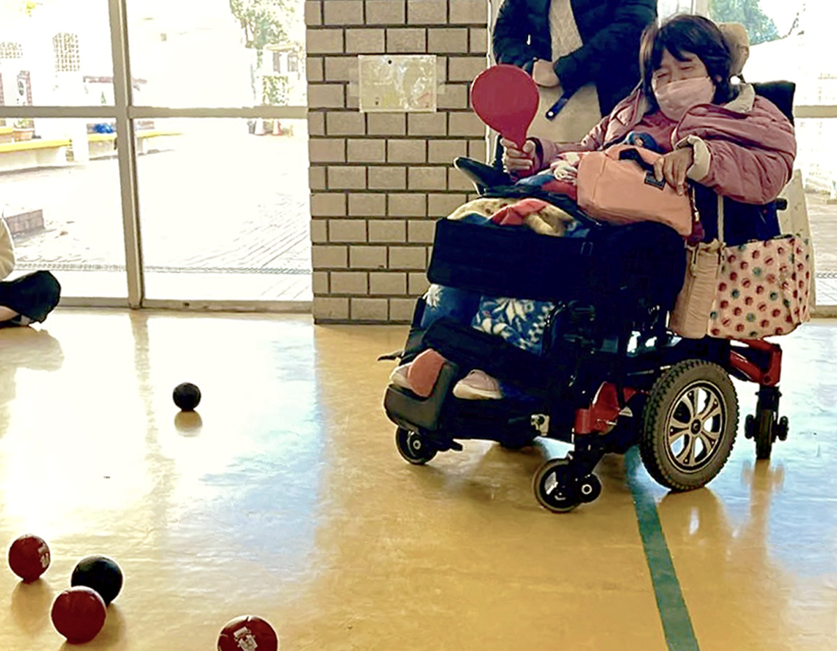 体育館のような場所で、車いすに乗った女性が床に転がっているボッチャの赤や青の球の位置をみながら審判をしている。