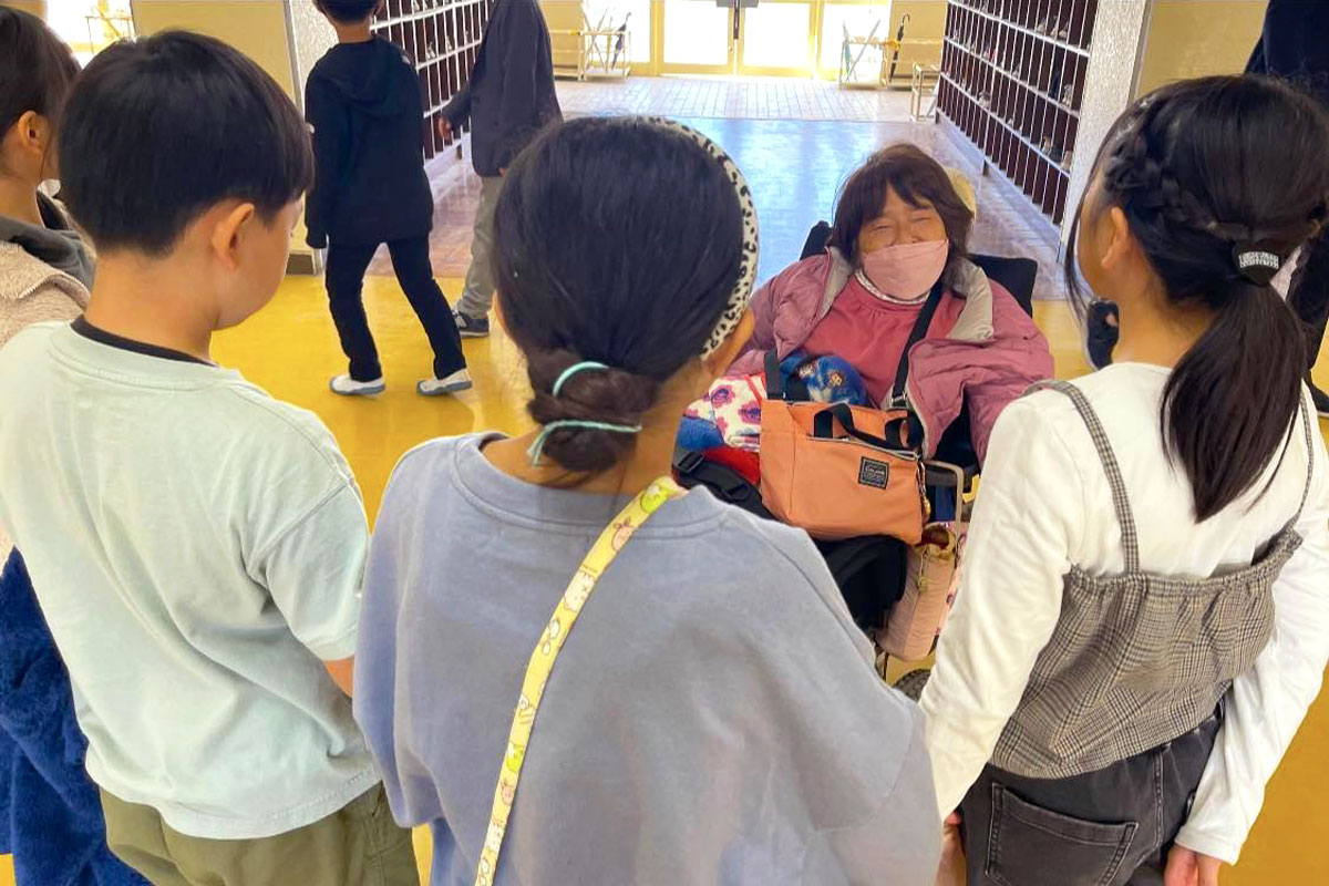 学校の下駄箱がならぶ昇降口のフロアで、子どもたちに囲まれる車いすに乗った女性。