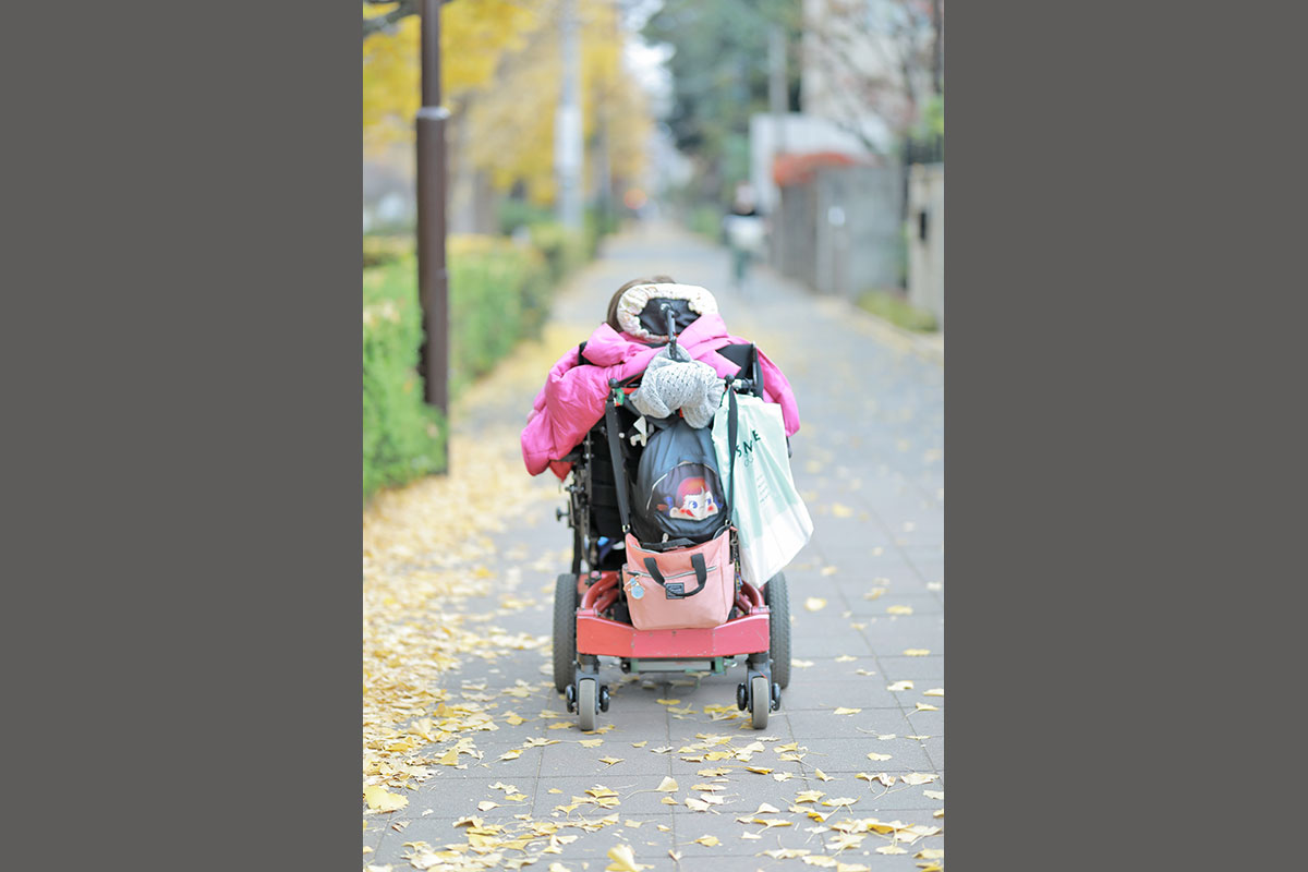 くにたちの大学通りの歩道がまっすぐに写っており、電動車いすに乗った女性の後ろ姿が写っている。