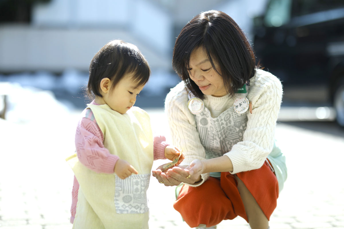 ２歳くらいのエプロンをした子が、かがんだ姿勢のおばちゃんの両手に乗せられた小さな紙を持ち上げ、見ている。