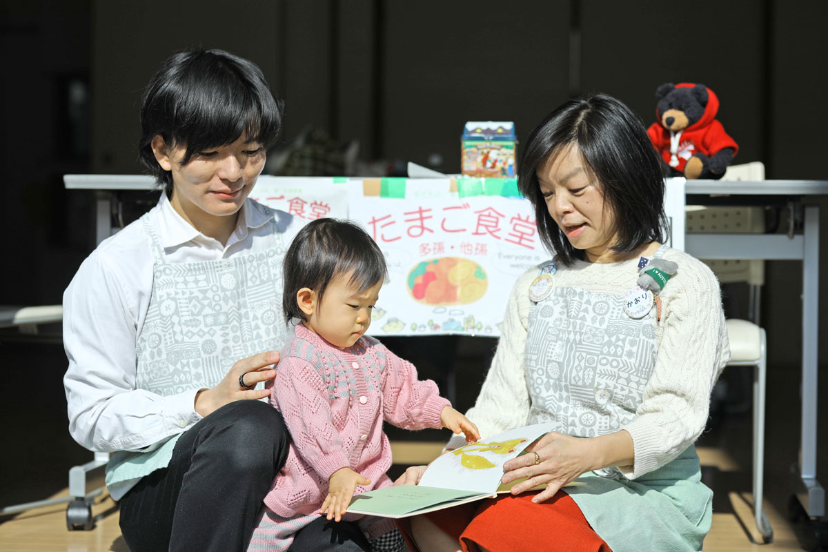 ２歳くらいの子に女性が絵本を手に持って読み聞かせをしている。