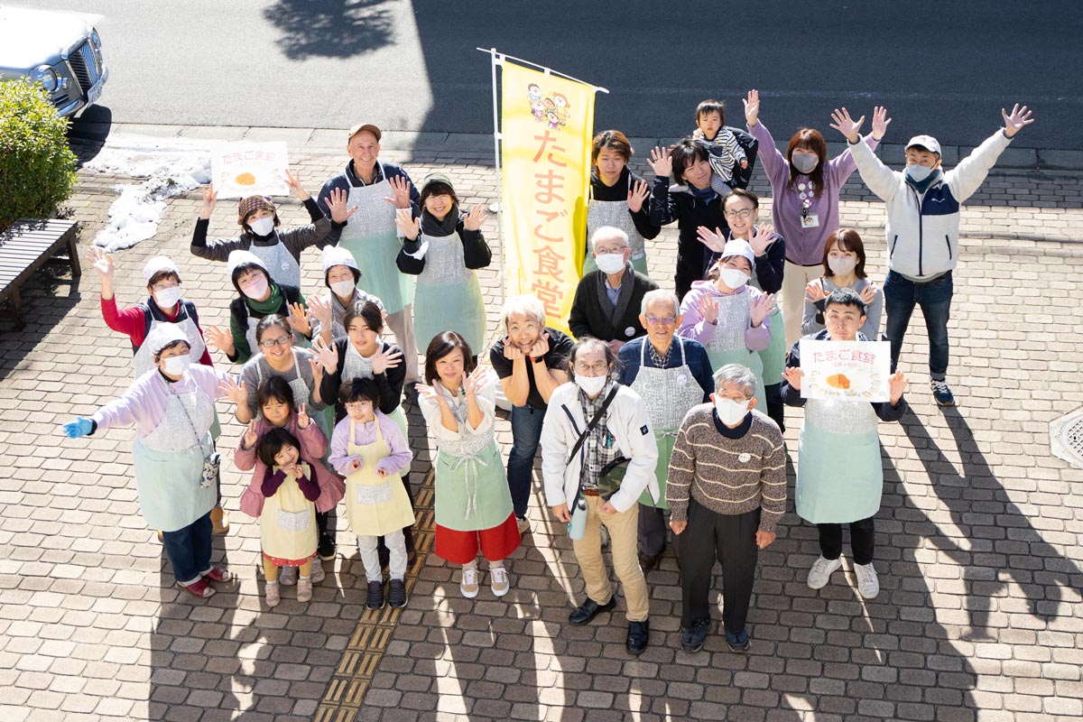 たまご食堂とかかれた黄色いのぼりを中心に30人ほどの人が思い思いにポーズをとって集合写真に写っている。