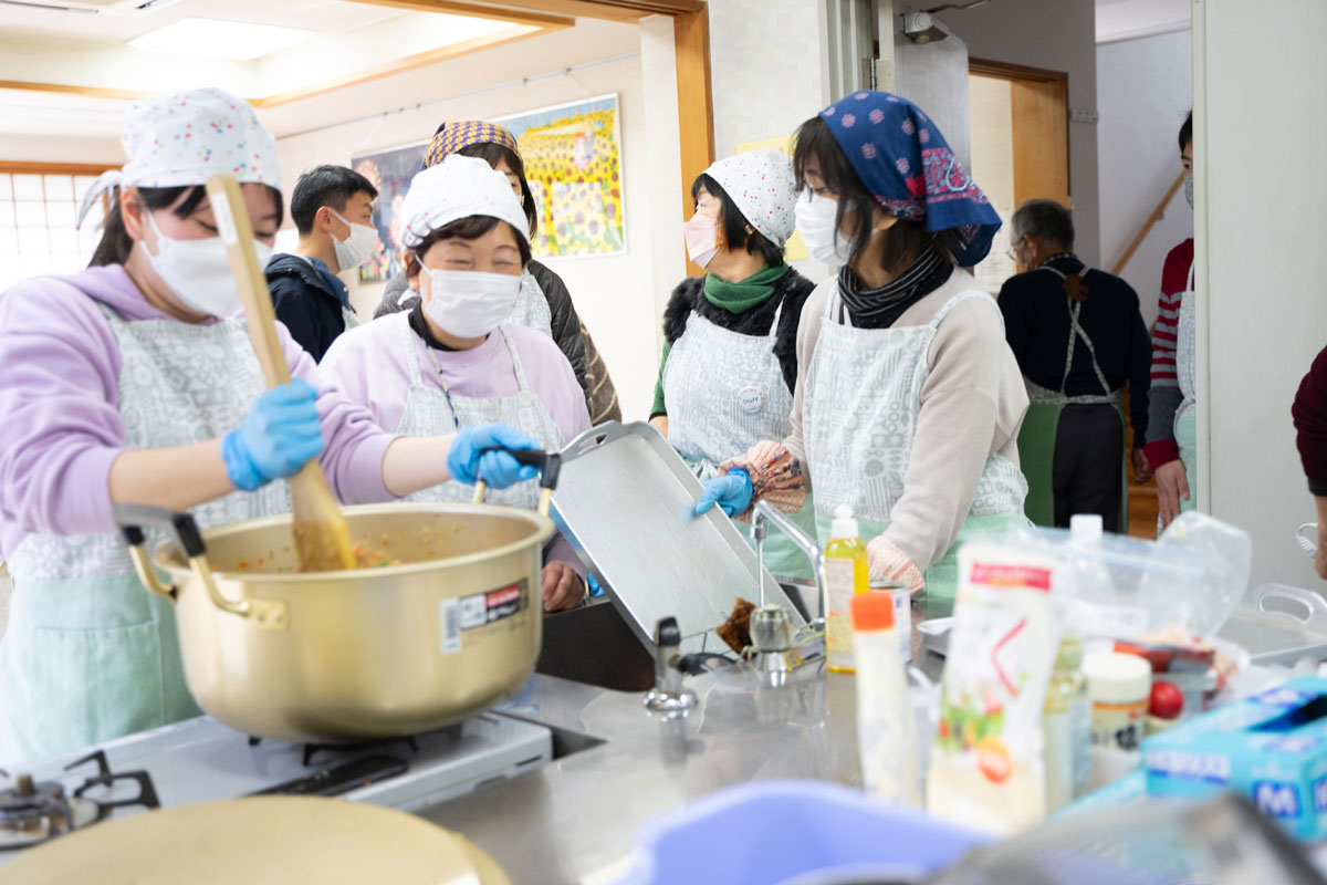 屋内で、エプロンと三角巾をしたみなさんが配膳の準備をしている。