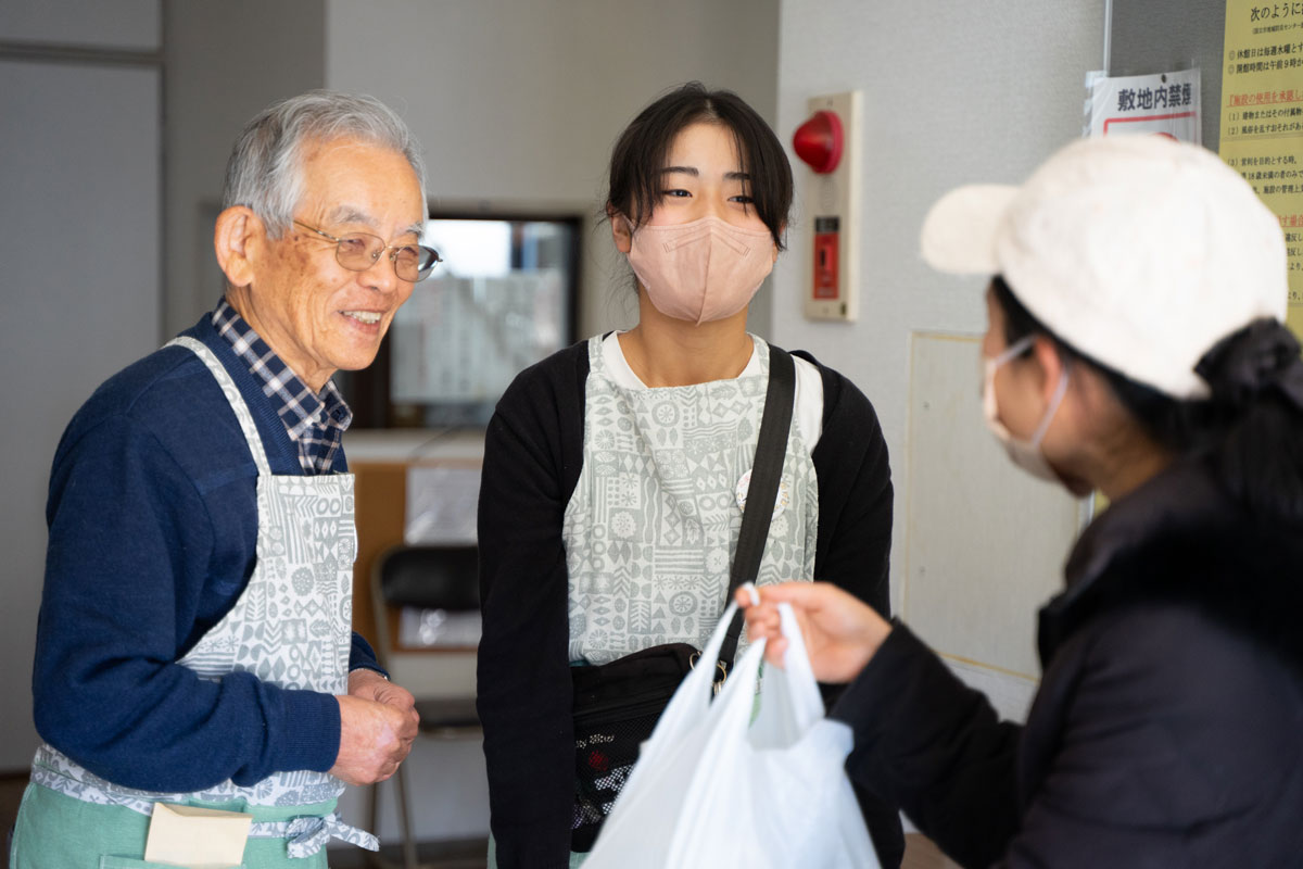 防災センターの入り口玄関で、来訪者とエプロンをした２人が談笑している。来訪者の方が白いビニール袋を持ち上げて持っているのでテイクアウトするのか差し入れかもしれない。