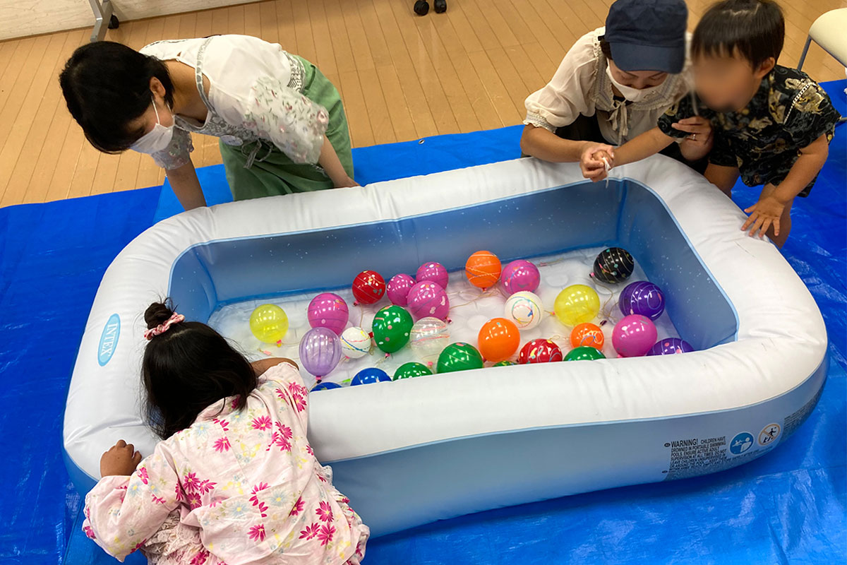 水風船のヨーヨー釣りに2組の親子が挑んでいる。小さな女の子と見守るおかあさん。もう一組は小さな男の子とおかあさんで、一緒にそーっと釣りあげている様子。