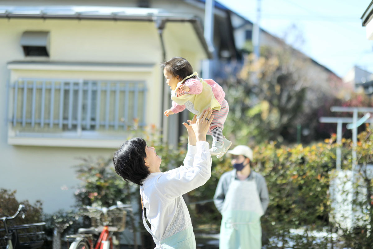 エプロンをした男性が小さい子をだきあげ、たかいたかいをしている。