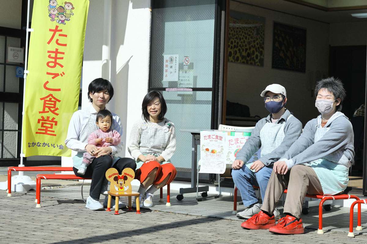 たまご食堂、なかなかいい会と書かれたのぼりの脇に低いベンチが並んでいる。こしかけて微笑んでいる運営スタッフのお兄さんとおばちゃん。お兄さんに抱きかかえられた小さな子は不思議そうな顔をしている。