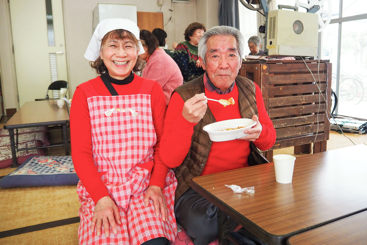 集会所に低めの長机が並び、囲むように座布団がしいてある。そこに赤いセーターにチェックのエプロンをして三角巾をかぶった笑顔の女性と、同じく赤いセーターを着てカレーを食べる男性が2人となりあって写っている。