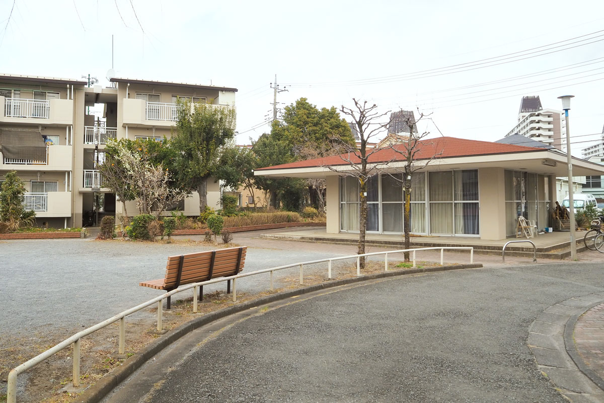 3階建て団地の手前に広場があり、ベンチも見える。その脇に赤茶色の屋根をした集会所が建っている。