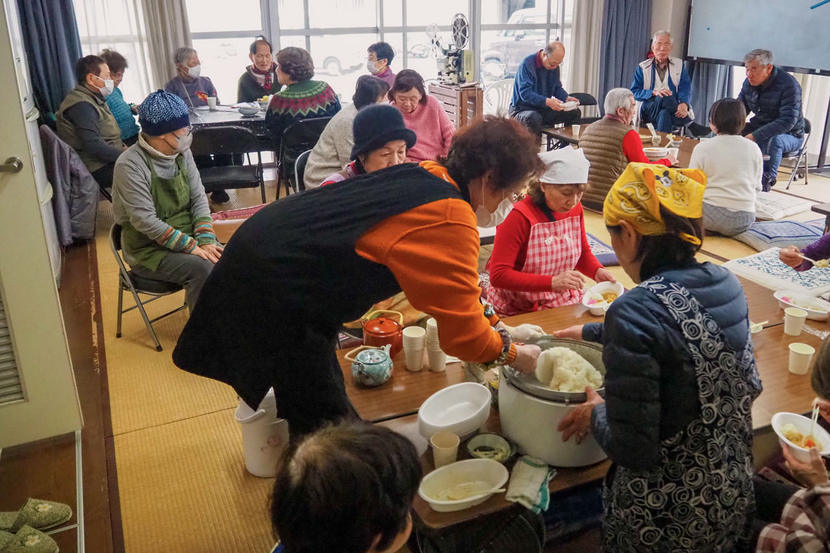 集会所の畳敷きの部屋にテーブルが５つくらいの島にわけて用意されていて、それぞれの卓を囲んでカレーを食べたり話をしている。手前の机では炊飯器のごはんをかき混ぜるエプロン姿の女性たちが写っている。