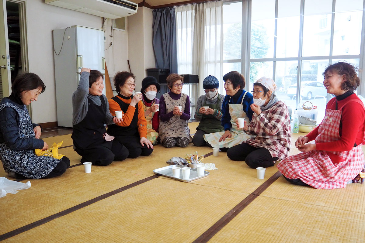 サッシのガラス戸に囲まれた和室で、周囲の光が差し込み明るい。畳の上では女性たちがあつまって、お茶を片手に談笑している。