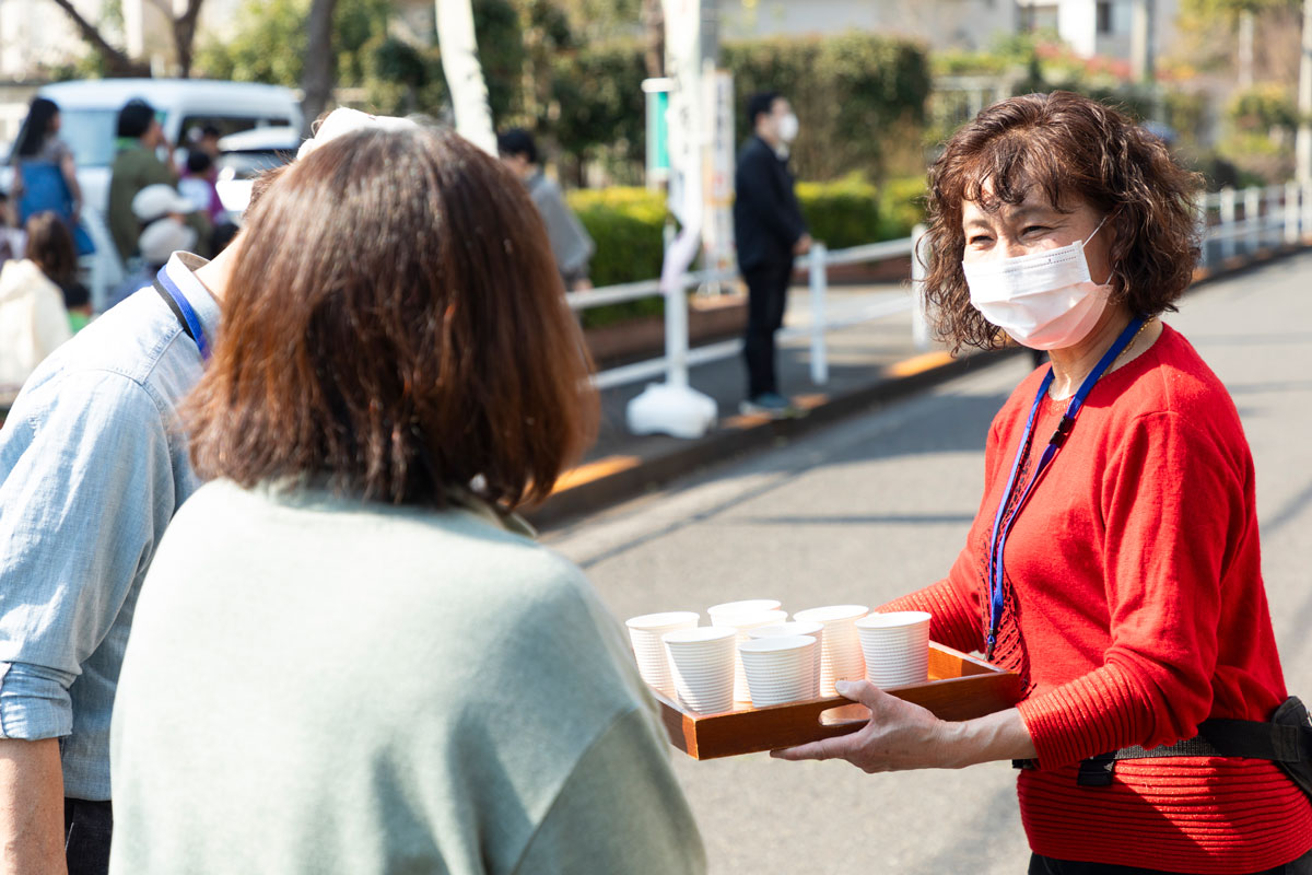 赤い服を着て、名札を下げた女性がコップの並んだお盆を手に立っている。笑顔で参加者に飲みものをすすめている様子。