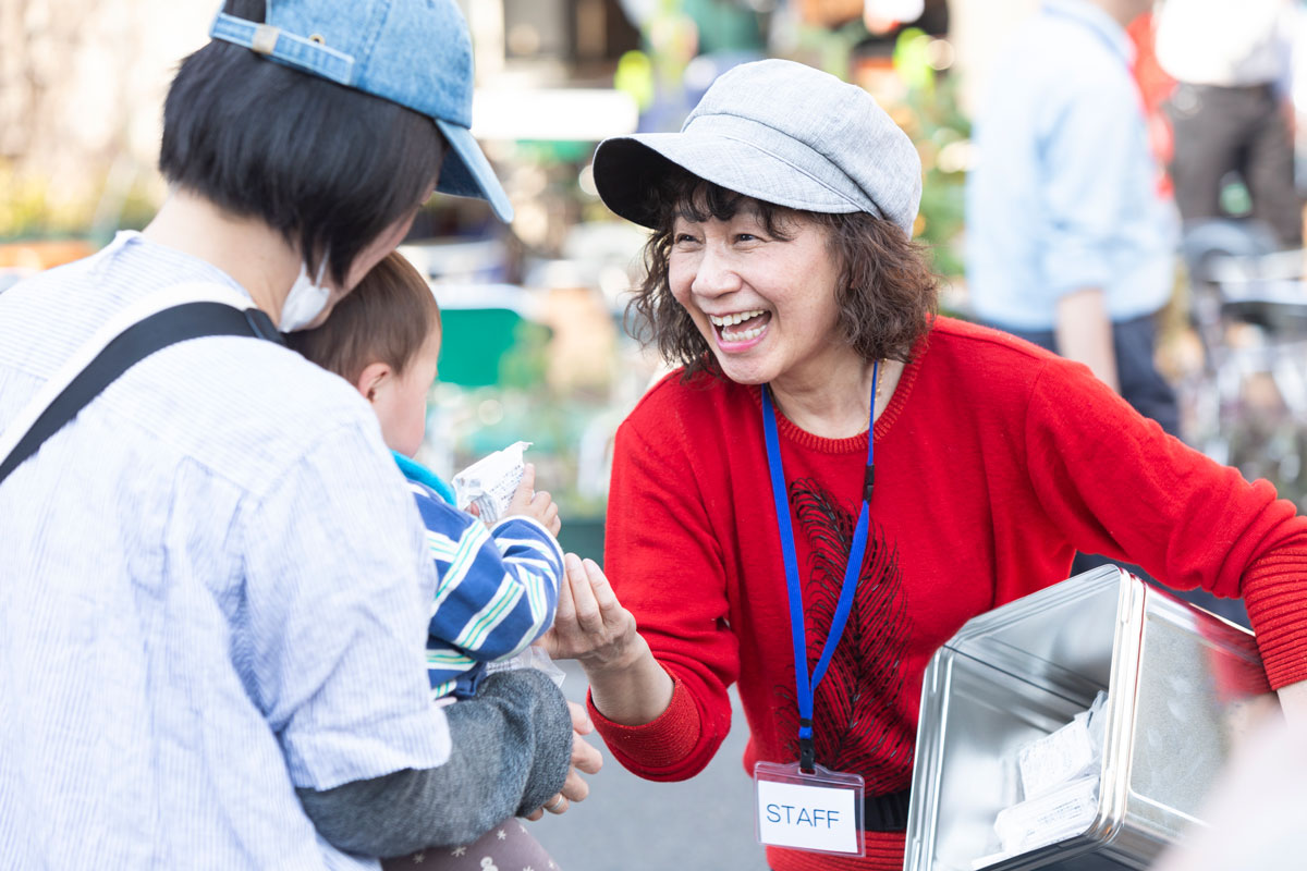 イベントスタッフの名札を下げ、参加者へ笑顔で景品を配っている女性。女性に抱えられた幼児がそれを受け取っている。